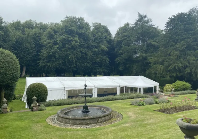 A large clearspan marquee of Melody Corporation set up in a lush garden with a fountain in the foreground and dense trees in the background.