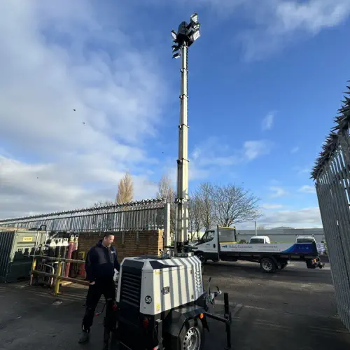 A man stands next to a Melody Corporation lighting tower extended high above in a fenced industrial area under a partly cloudy sky.