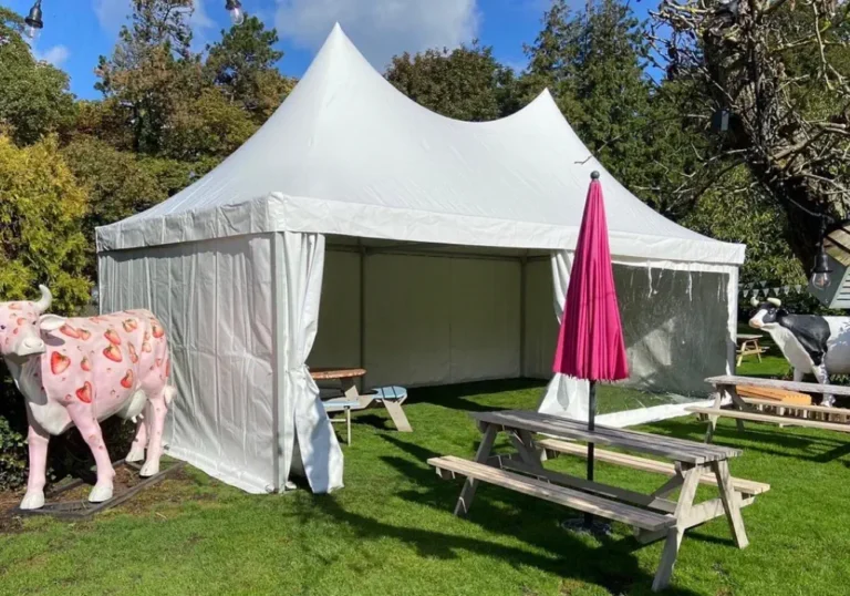 A large white pagoda marquee of Melody Corporation set up in a grassy area with picnic benches, a pink umbrella, and a decorative cow sculpture nearby.