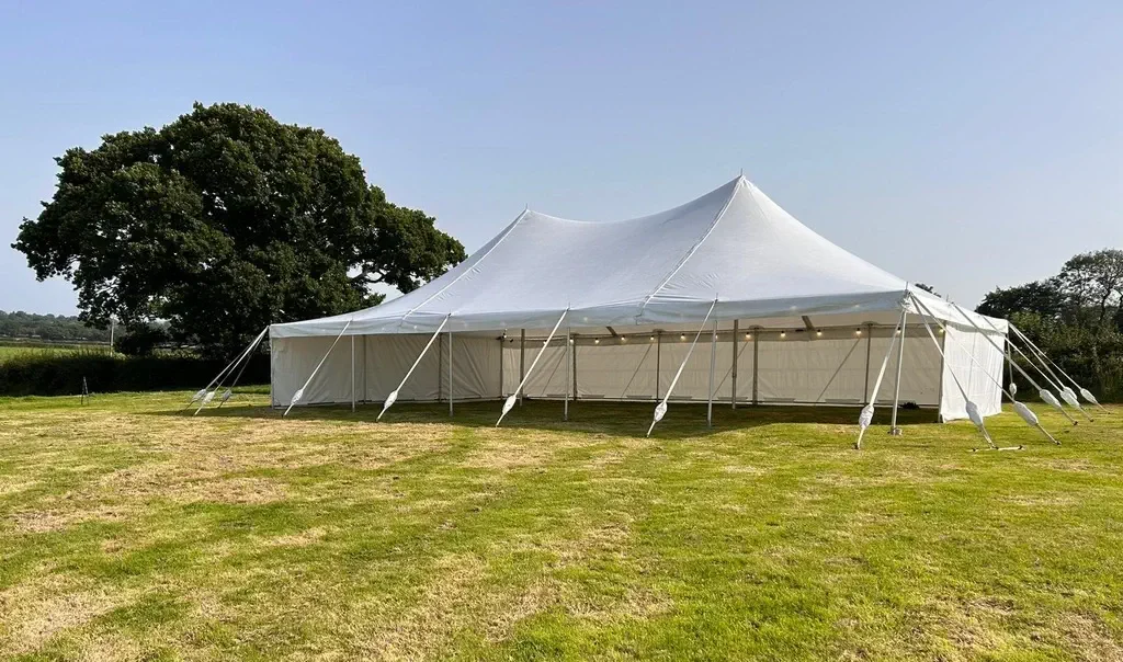 A large white traditional marquee is set up on a grassy field, secured with multiple ropes. Trees are visible in the background under a clear sky.