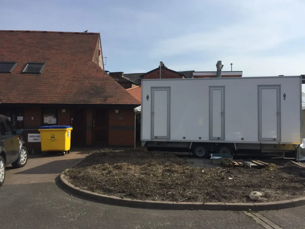 A white mobile unit is parked next to a building with sloped roofs, resembling a marquee. Nearby, a yellow bin and a black vehicle add to the scene. The foreground features a circular patch of dirt and mulch, complementing the surroundings like thoughtful accessories.