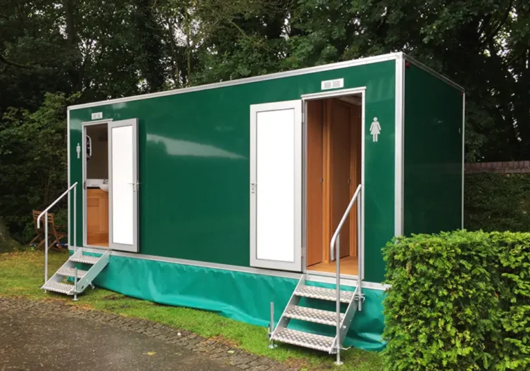 A green mobile restroom trailer with two open entry doors, one on the left marked for men and one on the right marked for women, is parked near a marquee surrounded by grass, a hedge, and trees.