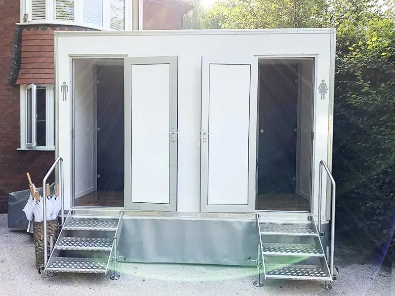 A portable toilet unit with two separate doors, one marked with a male symbol and the other with a female symbol, stands adjacent to a brick building. Each entrance features steps leading up and is complemented by marquee-style signage, blending essential accessories for convenience and style.