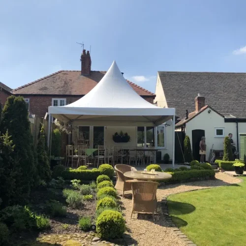 A landscaped backyard with a patio area covered by a white canopy, resembling elegant marquees. There are wicker furniture and a small path leading to a lawn. Two people stand near a shed and a house in the background, creating an inviting outdoor retreat.