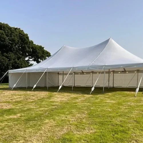 A large white traditional marquee is set up on a grassy field, secured with multiple ropes. Trees are visible in the background under a clear sky.
